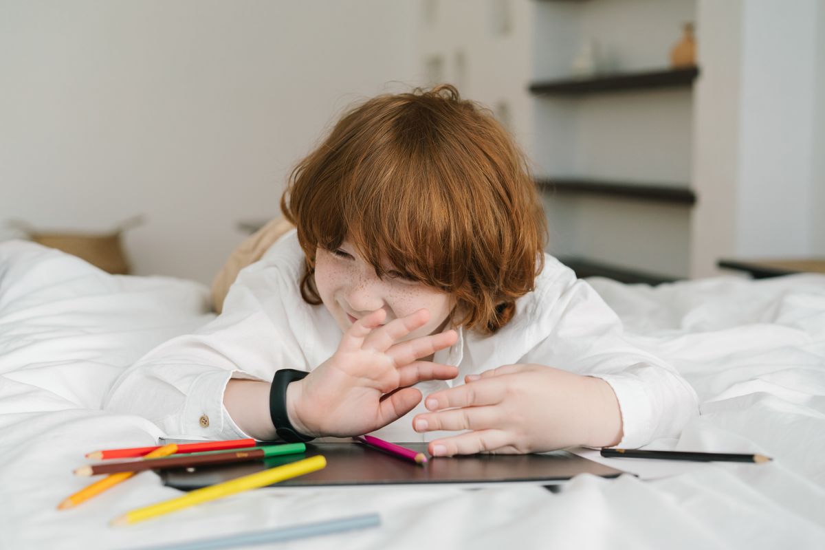 A young redheaded boy lying on a bed, playing with colored pencils indoors, expressing creativity.