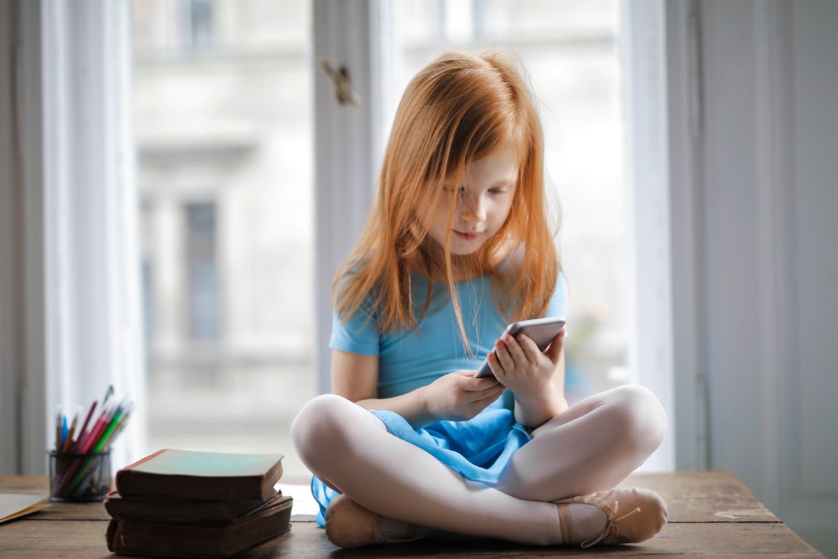 Red haired charming schoolgirl in blue dress browsing smartphone while sitting on rustic wooden table with legs crossed beside books against big window at home - Photo by Andrea Piacquadio on Pexels