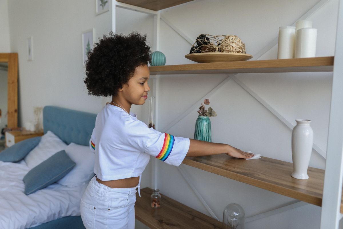 Charming African American girl wiping dust from wooden shelf of rack at home Charmantes afroamerikanisches Mädchen, das zu Hause Staub vom Holzregal des Regals wischt