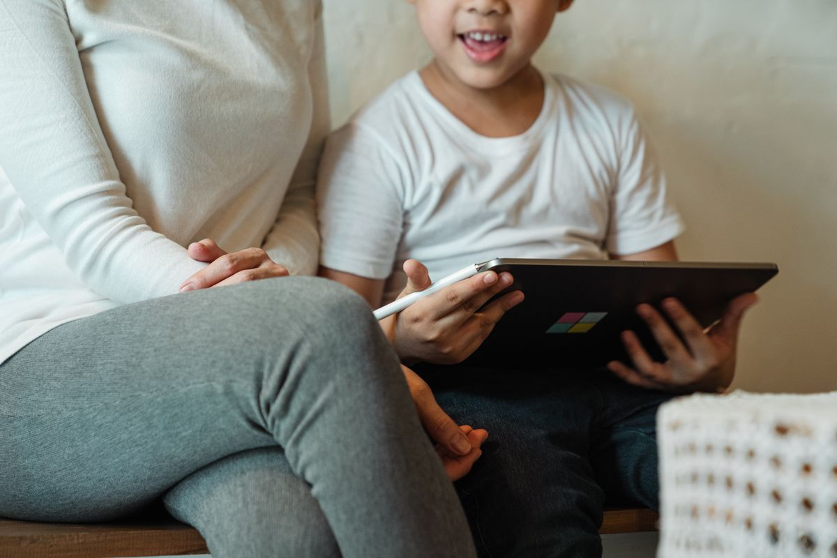 Exited crop schoolchild surfing internet on tablet while resting together with mother in domestic cloth at home during weekend