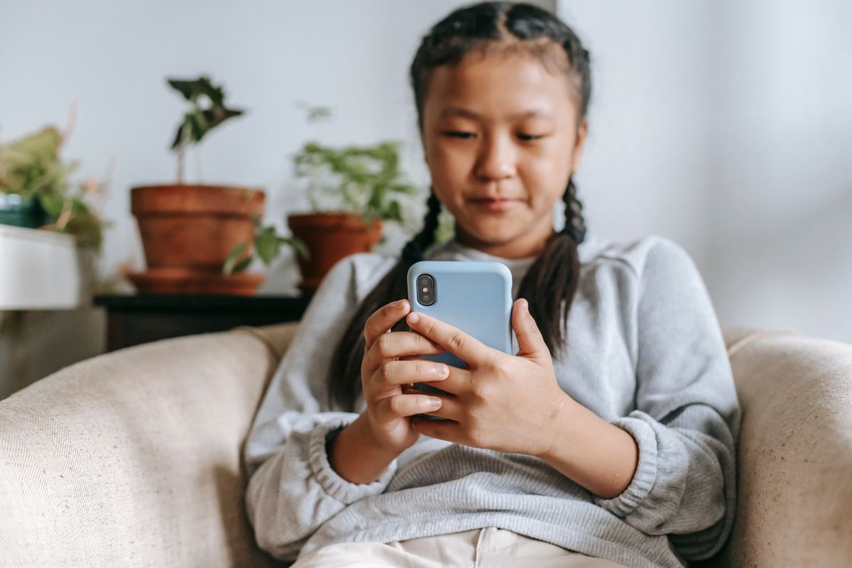 Focused Asian girl in casual clothes sitting on cozy armchair and browsing social media on smartphone