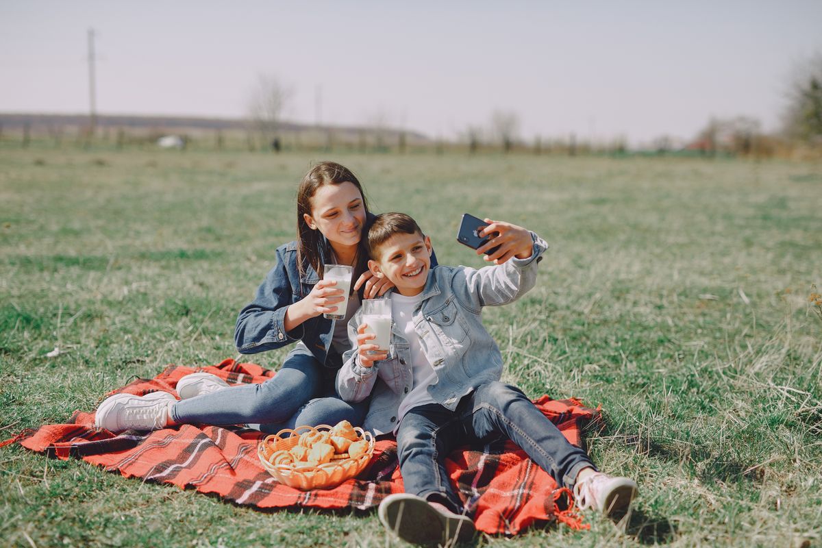 Smiling young boy and girl in casual outfit sitting together on checkered plaid with glass of milk and buns while taking selfie on mobile phone in park - Photo by Gustavo Fring on Pexels