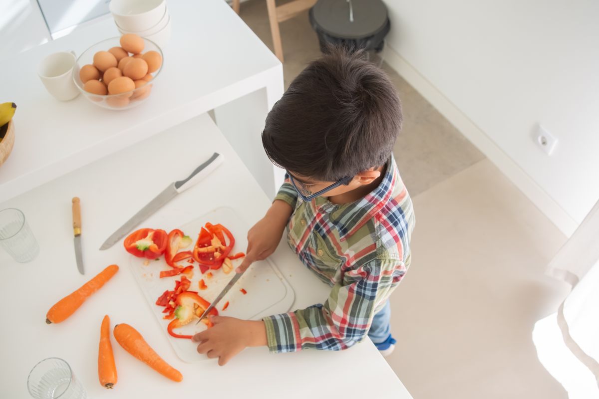 Ein Kind in einem karierten Hemd schneidet Gemüse auf einem Schneidebrett in einer modernen Küche. - Foto von Kampus Production auf Pexels