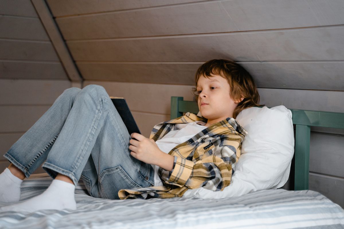 A young boy is lying on a bed reading a book in a cozy, wooden-themed bedroom. - Photo by MART  PRODUCTION on Pexels