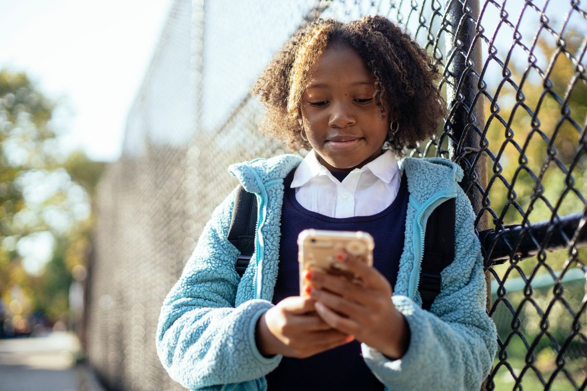 Colegiala afroamericana positiva con chaqueta azul navegando por teléfono móvil mientras está de pie cerca de una valla de alambre en un día soleado de otoño - Foto de Mary Taylor en Pexels