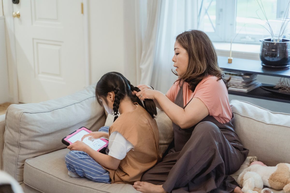 Photo by Nicola Barts on Pexels Eine Mutter flechtet die Haare ihrer Tochter, während sie im Wohnzimmer ein Tablet benutzt. - Foto von Nicola Barts auf Pexels