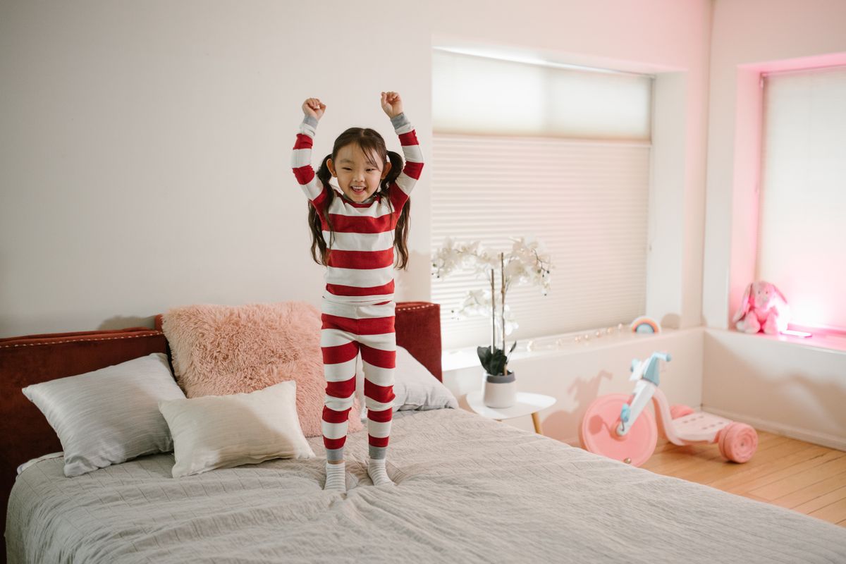 Ein fröhliches Kind im gestreiften Pyjama, das in einem hellen und gemütlichen Schlafzimmer auf ein Bett springt. - Foto von RDNE Stock-Projekt auf Pexels