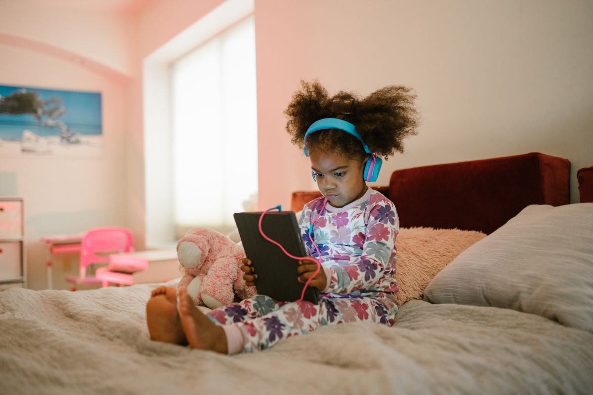 Una ragazza in pigiama si siede sul letto con le cuffie e usa un tablet. Ambiente domestico accogliente. - Foto del progetto RDNE Stock su Pexels