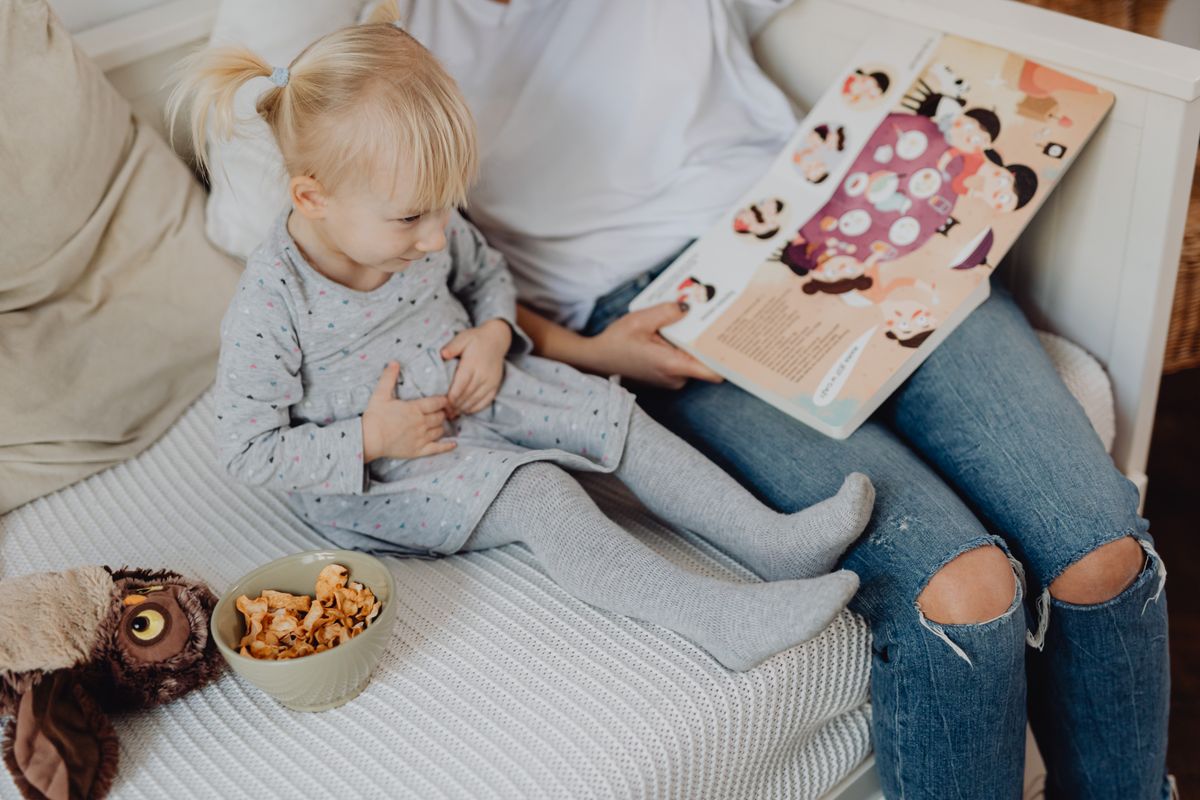 Mother reading a book to her daughter in a cozy bedroom setting with snacks nearby. - Photo by www.kaboompics.com on Pexels